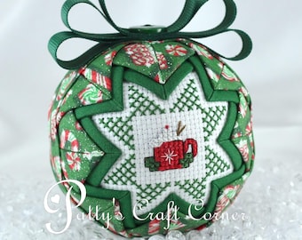 May include: A quilted fabric ornament featuring a green and red candy cane pattern with a central white star patch displaying a cross-stitched red mug of hot cocoa. The ornament is topped with a dark green ribbon loop for hanging.
