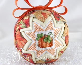 May include: A spherical fabric ornament featuring a quilted star pattern with autumn-themed leaf prints in orange, red, and yellow. A central cross-stitched panel displays a small orange pumpkin with green leaves. An orange ribbon loop sits on top.
