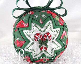 May include: A quilted fabric Christmas ornament featuring a green and red candy cane pattern with a central white cross-stitched panel displaying two candy canes tied with a green bow. The ornament is topped with a dark green ribbon loop.