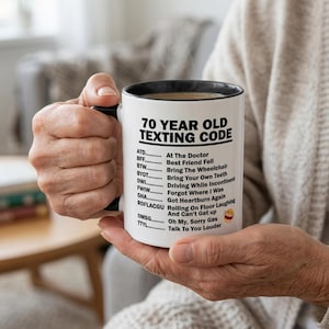 May include: A white ceramic mug with a black handle and rim, featuring the black printed text 70 YEAR OLD TEXTING CODE followed by a list of humorous acronyms and their meanings, ending with a laughing emoji with tears of joy.