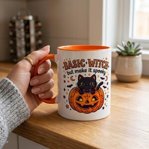 May include: A white ceramic mug with an orange handle and interior rim. The front features a graphic of a black cat sitting inside a carved orange pumpkin. The text reads "BASIC WITCH but make it spooky" surrounded by small stars, moons, and bats.