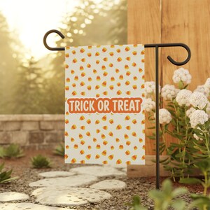 May include: A rectangular garden flag featuring a repeating pattern of orange, yellow, and white candy corn shapes on a cream background. The center of the flag displays the orange text TRICK OR TREAT in a bold, rounded, sans-serif font.