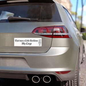 May include: A rectangular white bumper sticker applied to the rear of a grey car. The sticker features black text in a gothic font that reads, "That was a Little Reckless My Liege," accompanied by a small black line drawing of a jester figure.