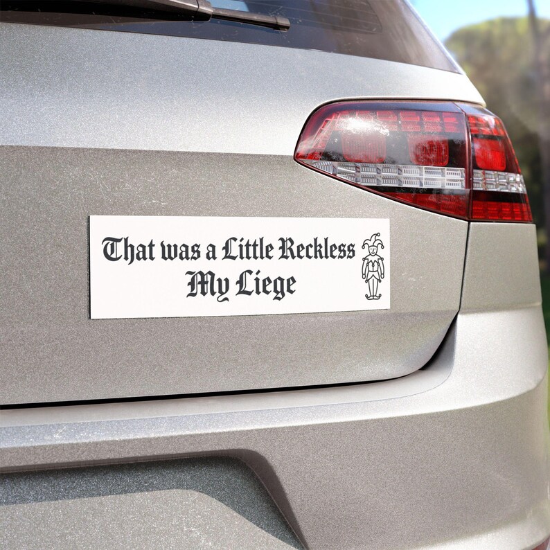 May include: A rectangular white bumper sticker with black text that reads "That was a Little Reckless My Liege" in a gothic-style font. To the right of the text is a small, simple black line drawing of a jester wearing a traditional cap.