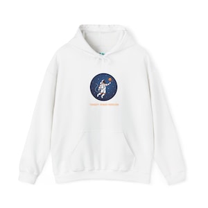 May include: A white hooded sweatshirt featuring a circular graphic on the chest. The graphic depicts an astronaut floating in dark blue space, reaching towards a small orange planet. Below the circle, the text reads CURIOSITY: PRIMARY PROPULSION.