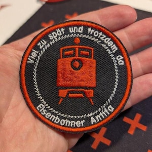 Könnte beinhalten: Ein kreisförmiger gestickter Aufnäher mit schwarzem Hintergrund und orangefarbenem Rand. In der Mitte befindet sich ein orangefarbenes Lokomotivsymbol. Der Text lautet oben "Viel zu spät und trotzdem da" und unten "Eisenbahner Antifa" in Weiß.