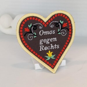 Könnte beinhalten: Ein herzförmiger Aufnäher mit dunkelbraunem Hintergrund und cremefarbenem Rand. Der Text Omas gegen Rechts ist in Weiß gestickt, umgeben von roten Zierpunkten, schwarzen Schnörkeln sowie kleinen roten und gelben floralen Akzenten.