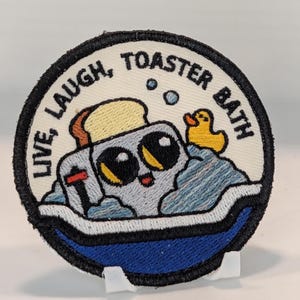 Könnte beinhalten: Ein runder, bestickter Aufnäher mit schwarzem Rand auf cremefarbenem Hintergrund. Er zeigt einen grauen Toaster mit großen Augen und einer Scheibe Toast in blauem Wasser mit Blasen und einer gelben Quietscheente. Der Text lautet: LIVE, LAUGH, TOASTER BATH.