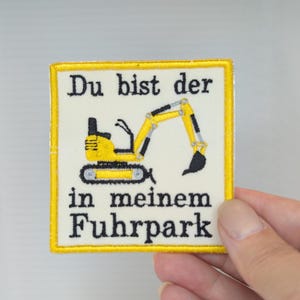 Könnte beinhalten: Ein quadratischer Aufnäher mit cremefarbenem Hintergrund und gelbem Rand. Er zeigt eine gelbe und schwarze Bagger-Grafik sowie den in Schwarz gestickten Text "Du bist der in meinem Fuhrpark", der mittig auf der Stoffoberfläche platziert ist.