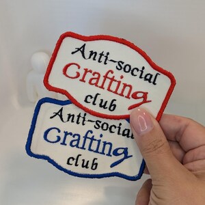 Könnte beinhalten: Zwei rechteckige bestickte Aufnäher mit cremefarbenem Stoffuntergrund. Der obere Aufnäher hat einen roten Rand und den Text "Anti-social Grafting club" in Schwarz und Rot. Der untere Aufnäher hat einen blauen Rand und denselben Text in Schwarz und Blau.