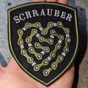 Könnte beinhalten: Ein schildförmiger Aufnäher mit schwarzem Hintergrund und grünem Rand. Der Schriftzug SCHRAUBER ist in Weiß im oberen Bereich gestickt. Darunter bildet eine Reihe von miteinander verbundenen olivgrünen Fahrradkettengliedern eine Herzform.