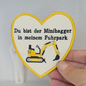 Könnte beinhalten: Ein herzförmiger Stickaufnäher mit cremefarbenem Hintergrund und gelbem Rand. Der Aufnäher zeigt eine gelb-schwarze Bagger-Illustration und den schwarzen Sticktext: Du bist der Minibagger in meinem Fuhrpark.