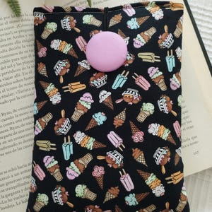 Puede incluir: Una funda de tela rectangular con fondo negro y un patrón repetitivo de conos de helado y polos coloridos en tonos rosa, verde, azul y marrón. La funda se asegura con un único botón de tela de color morado claro y liso.