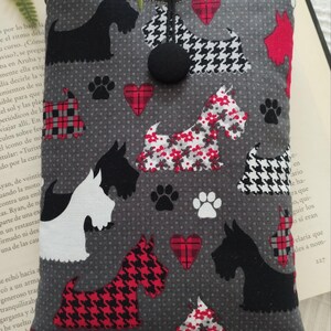 Puede incluir: Una funda rectangular de tela en gris oscuro con un patrón repetitivo de siluetas de terrier escocés, huellas de patas y corazones de cuadros rojos. La funda cuenta con un cierre de botón negro en la parte superior central.