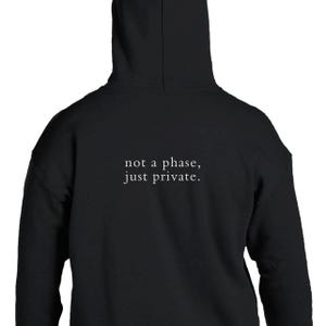 Könnte beinhalten: Ein schwarzer Kapuzenpullover mit dem weißen Text "not a phase, just private." auf der Rückseite. Das Kleidungsstück verfügt über eine Kapuze und lange Ärmel und zeigt ein schlichtes Design mit klarer, zentrierter Typografie im oberen Rückenbereich.