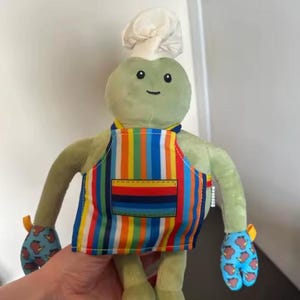 May include: A plush toy shaped like a green creature wearing a white chef hat, a colourful vertical striped apron with a front pocket, and blue oven mitts featuring a repeating pattern of small brown kiwi birds on each hand.