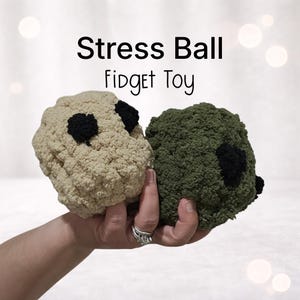 May include: Two textured stress balls, one beige and one olive green, each with two black accents. The balls are held in a hand. Text above reads "Stress Ball Fidget Toy".