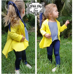 May include: A young girl wearing a yellow cardigan with a blue and white striped shirt underneath. She is also wearing navy blue leggings and white shoes. The cardigan has a long, flowing back.