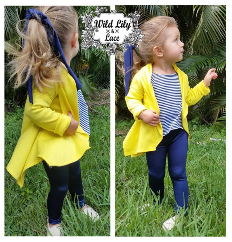 May include: A young girl wearing a yellow cardigan with a blue and white striped shirt underneath. She is also wearing navy blue leggings and white shoes. The cardigan has a long, flowing back.