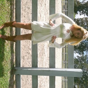 May include: A woman wearing a white, lace-trimmed dress with bell sleeves and brown cowboy boots. She is standing in front of a white wooden fence.