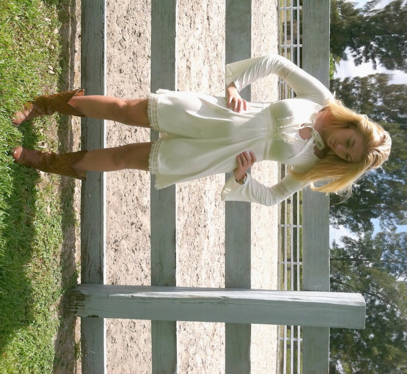 May include: A woman wearing a white, lace-trimmed dress with bell sleeves and brown cowboy boots. She is standing in front of a white wooden fence.