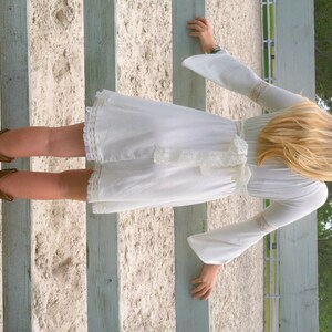 May include: A woman wearing a white dress with a lace trim and a bow at the waist. She is standing in front of a wooden fence and wearing brown cowboy boots.