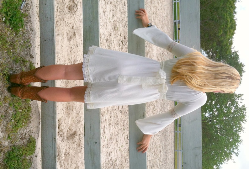 May include: A woman wearing a white dress with a lace trim and a bow at the waist. She is standing in front of a wooden fence and wearing brown cowboy boots.