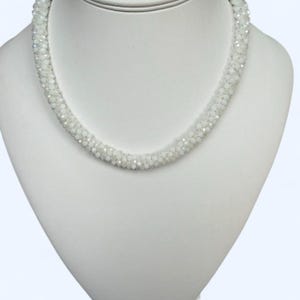 May include: A necklace composed of numerous small, translucent white beads with an iridescent finish. The beads are densely packed together to form a thick, cylindrical strand that curves in a circular shape around the neck display form.