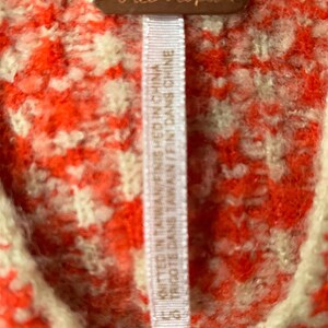 Może przedstawiać: Biała metka z czarnym tekstem, na kt&oacute;rej widnieje napis "Free People" oraz "KNITTED IN TAIWAN/FINISHED IN CHINA" i "TRIGOTE DANS TAWAIN/FINT DANS CHINE", przymocowana do czerwono-białego swetra.