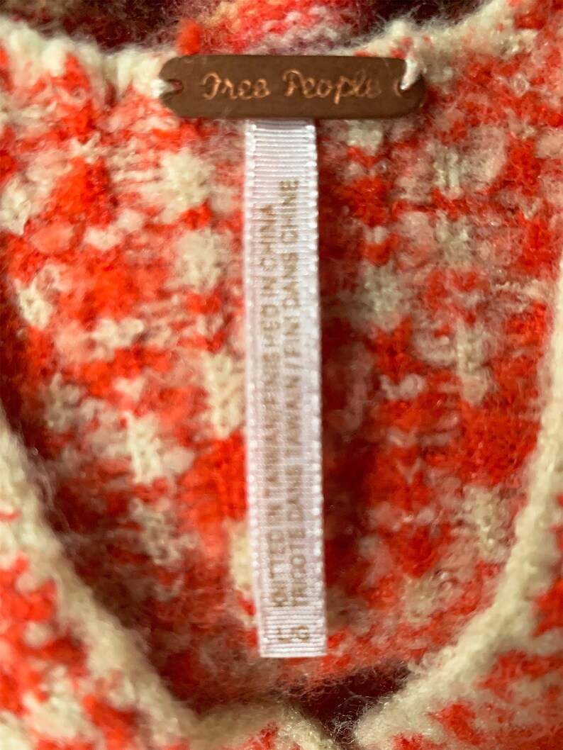 Może przedstawiać: Biała metka z czarnym tekstem, na kt&oacute;rej widnieje napis "Free People" oraz "KNITTED IN TAIWAN/FINISHED IN CHINA" i "TRIGOTE DANS TAWAIN/FINT DANS CHINE", przymocowana do czerwono-białego swetra.