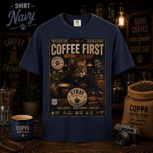 May include: A navy blue short-sleeve t-shirt featuring a graphic print of a cat wearing a beanie and hoodie. The text reads COFFEE FIRST THEN CATNAPS, COPPA COFFEE CLUB, STRAY COFFEE CLUB, and GOOD COFFEE GOOD COMPANY GOOD CAT, along with various icons.
