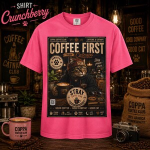 May include: A bright pink short-sleeve t-shirt featuring a graphic print of a cat wearing a beanie and hoodie. The text reads COFFEE FIRST THEN CATNAPS, COPPA COFFEE CLUB, and STRAY COFFEE CLUB, along with various coffee-themed icons and phrases.