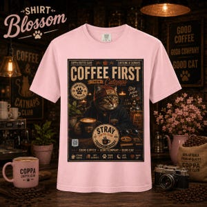 May include: A light pink t-shirt featuring a graphic print of a cat wearing a beanie and hoodie, sitting at a table with a coffee mug. The text reads COFFEE FIRST THEN Catnaps, COPPA COFFEE CLUB, CAFFEINE & CATNAPS, PAWSITIVE LIFESTYLE, and STRAY COFFEE CLUB.