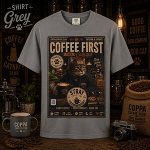 May include: A grey t-shirt featuring a graphic print of a cat wearing a beanie and hoodie, sitting at a table with a coffee mug. The text reads COFFEE FIRST THEN Catnaps, COPPA COFFEE CLUB, STRAY COFFEE CLUB, and other coffee-related phrases.
