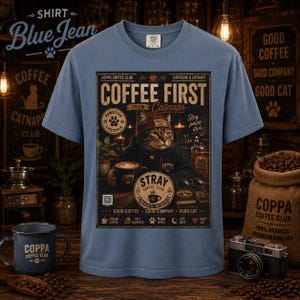 May include: A blue short-sleeve t-shirt featuring a graphic print of a cat wearing a beanie and hoodie. The text reads COFFEE FIRST THEN Catnaps, COPPA COFFEE CLUB, and STRAY COFFEE CLUB. The design includes coffee-related icons and phrases.