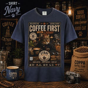 May include: A navy blue t-shirt featuring a graphic print of a cat wearing a beanie and hoodie, holding a coffee mug. The text reads COFFEE FIRST THEN CATNAPS, PAWSITIVE LIFESTYLE, STRAY COFFEE CLUB, and GOOD COFFEE GOOD COMPANY GOOD CAT.