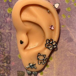 May include: A silicone ear model featuring multiple silver-toned piercings, including cat ear studs, a pink crystal stud, a heart stud, a paw print charm, a cat silhouette charm, and a small metallic frog figure at the bottom of the earlobe.