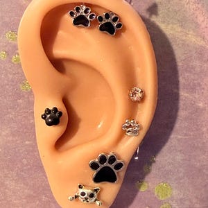 May include: A silicone ear model displaying multiple piercings featuring silver-tone jewelry with black enamel paw prints, a small cat face charm, a clear round gemstone stud, and two small pink conical studs at the top of the ear cartilage.