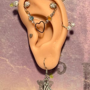 May include: A silver-toned ear cartilage piercing set featuring a chain with a central heart charm, blue and orange gemstone accents, and crystal-studded balls. Includes a small heart stud, a hoop with crystal balls, and a finger heart dangle earring.