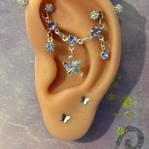 May include: An industrial barbell earring with a silver-tone chain featuring light purple crystals and a central butterfly charm. The jewelry includes two additional small butterfly stud earrings positioned on the earlobe, all set in silver-tone metal.