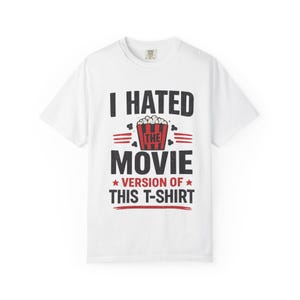 Può includere: Una t-shirt bianca a maniche corte con un design grafico composto da testo nero e rosso che recita I HATED THE MOVIE VERSION OF THIS T-SHIRT. Il design include un'illustrazione centrale di un secchiello per popcorn a righe rosse e nere.