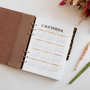 Peut inclure: Un classeur à la texture suédée marron avec un calendrier 2020, une règle marron translucide et des onglets noirs. Deux stylos sont présents : un à motif floral et un vert olive uni. La page du calendrier affiche le texte CALENDRIER.