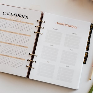 Op de afbeelding: Een open ringbandplanner met een 2021-kalender op de linkerpagina en een verjaardagstracker met de titel Anniversaires op de rechterpagina. De planner heeft zwarte tabbladen en wordt vergezeld door twee pennen met bloemmotieven en groene ontwerpen.