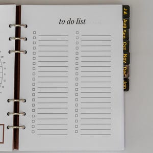 Puede incluir: Una página de agenda blanca con el encabezado to do list, que presenta dos columnas de líneas horizontales con casillas de verificación cuadradas. La página está encuadernada con un mecanismo de anillas metálicas y pestañas mensuales.