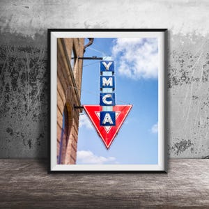 May include: Framed print of a vintage YMCA sign. The sign is composed of blue and white rectangular panels spelling "YMCA" above a red and white arrow. The sign is mounted on a brick building against a blue sky.