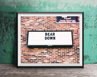 May include: Framed print featuring a sign that reads "BEAR DOWN" in bold white letters. The sign is mounted on a brick wall. The print is in a black frame and is set against a green and wood-grain background.