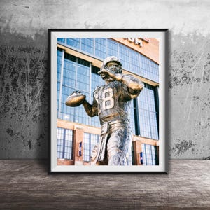 May include: A bronze statue of a football player in a throwing pose, wearing a jersey with the number 18. The statue is in front of a large building with glass windows.