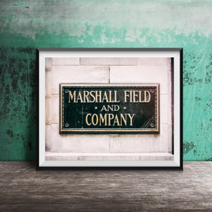 Op de afbeelding: Een rechthoekig zwart bord met gouden letters met de tekst MARSHALL FIELD AND COMPANY, gemonteerd op een lichtgekleurde stenen muur. Het bord is ingesloten in een dunne zwarte lijst en heeft een decoratieve rand met kleine ronde accenten.