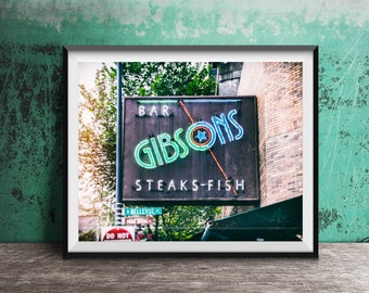 May include: A framed wall art print featuring a rectangular sign for Gibsons Bar Steaks-Fish with glowing neon lettering in green and blue. The sign includes a central star graphic and a diagonal orange neon line, set against a dark background.