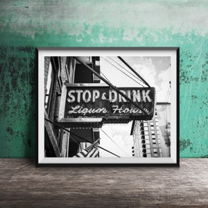 Op de afbeelding: Zwart-witfoto van een vintage neon-bord met de tekst "Stop & Drink Liquor House" dat boven een straat in een stad hangt. Het bord is met kettingen aan een gebouw bevestigd.